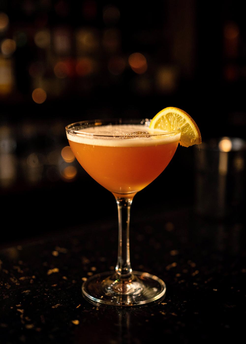 Amber craft cocktail in a coupe glass on a dark bar counter